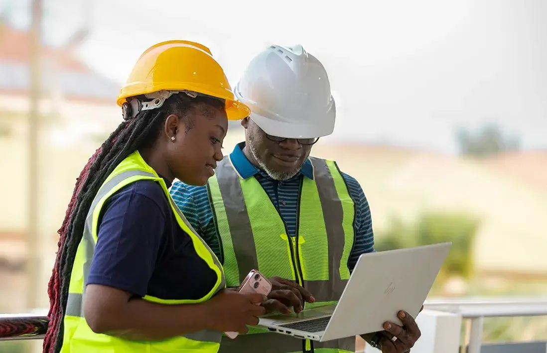 Two property professionals in hard hats reviewing plans on a laptop at a building site